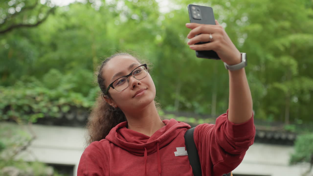 Mulher birracial tirando selfie sob as árvores, telefone levantado, poses animadas e sorriso sutil, luz do sol filtrando pelas folhas, moletom vermelho e óculos, vibe de criadora animada, compondo fotos.
