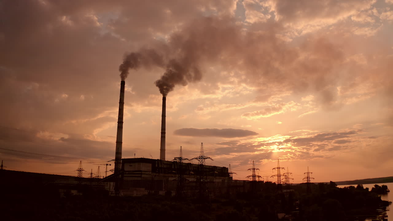 Harmful industrial area near the river. Power plant with smoking pipes pollute the atmosphere. Power station and factory at sunset. Industrial district. Smoke pipes against the evening sky.