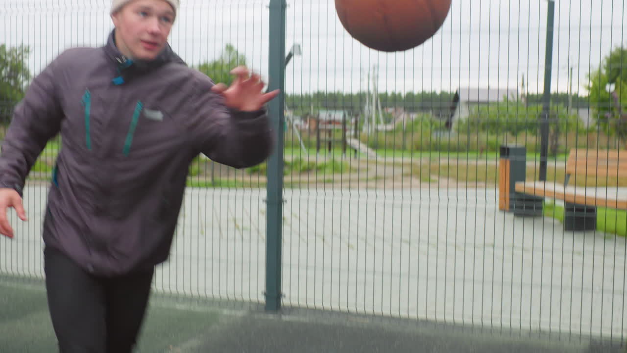 white male dribbling basketball outdoors wearing beanie and jacket, bouncing ball across fenced asphalt court near apartment building, focused expression as shot arcs toward rim and backboard, cool