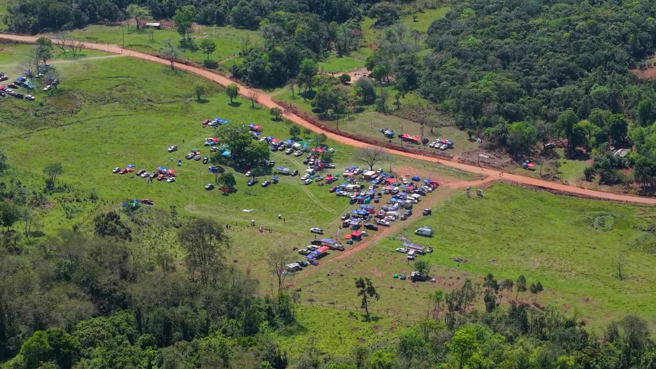 Midlertidig campingplads med telte, parkerede køretøjer og campere spreder sig ud over grønt landskab ved siden af en landlig grusvej i Nueva Alborada, Itapua, Paraguay, Sydamerika, droneoversigtsbillede