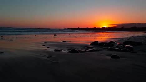 Low-dolly-shot-of-pebbles-at-beach-during-sunset-with-sunlight-reflecting-beautifully-on-the-sand-at-San-Buenaventura-State-Beach-in-Ventura,-California,-USA