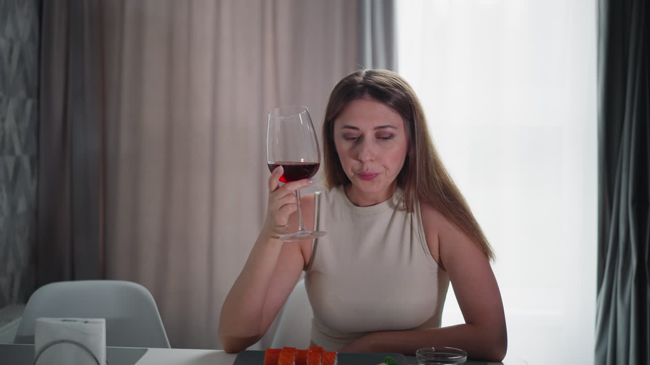 Young woman in sleeveless outfit drinks red wine at dining table with fresh fruit and napkin in front, soft natural light through curtains, casual indoor setting