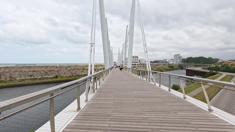 a steady forward-moving camera traverses a contemporary pedestrian bridge over a canal in dunkirk, france, under overcast daylight with soft, diffuse lighting