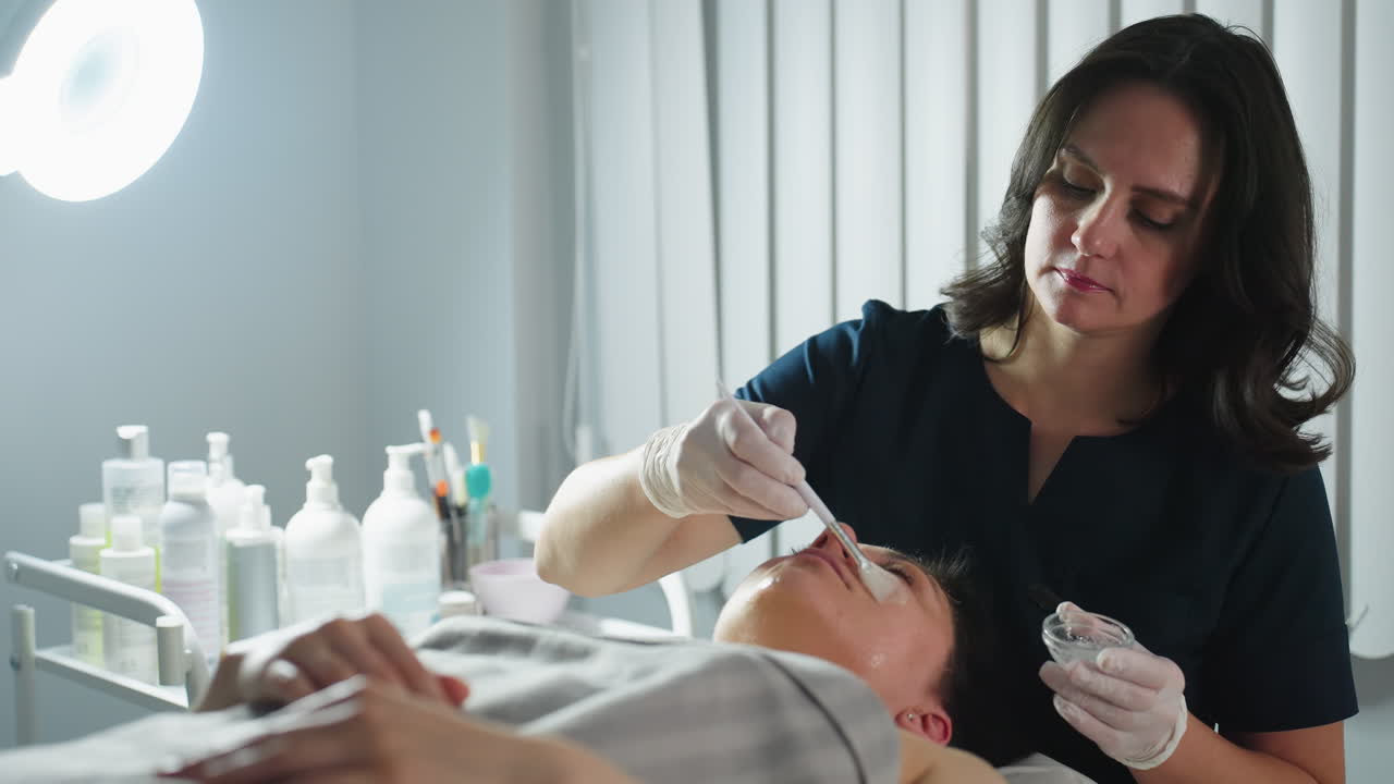 Beautician in gloves dips brush into clear gel then smooths cleansing mask in gentle circular strokes across closed eyes cheeks and forehead of relaxed client lying on spa bed for deep skin care
