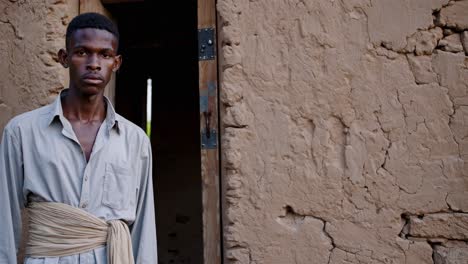 young african man wearing traditional clothes standing near mud house entrance, embodying rural lifestyle with quiet dignity and cultural authenticity