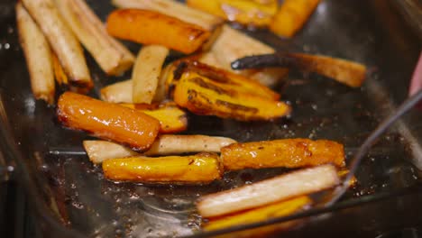 chef with spoon moving roast carrots and parsnips around on glass tray to cover in olive oil 4k