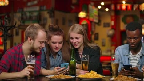 a man and two girls are sitting in a bar and looking at the phone screen while in the company of friends.