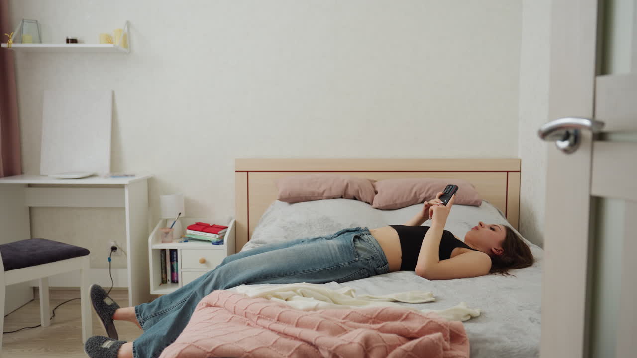 Young Woman Lying On Bed Scrolling Smartphone In Jeans And Black Top Under Pink Blanket, Bedside Shelf With Books And Lamp, Partially Open Door Framing Scene, Shoes At Foot Of Bed, Soft Daylight