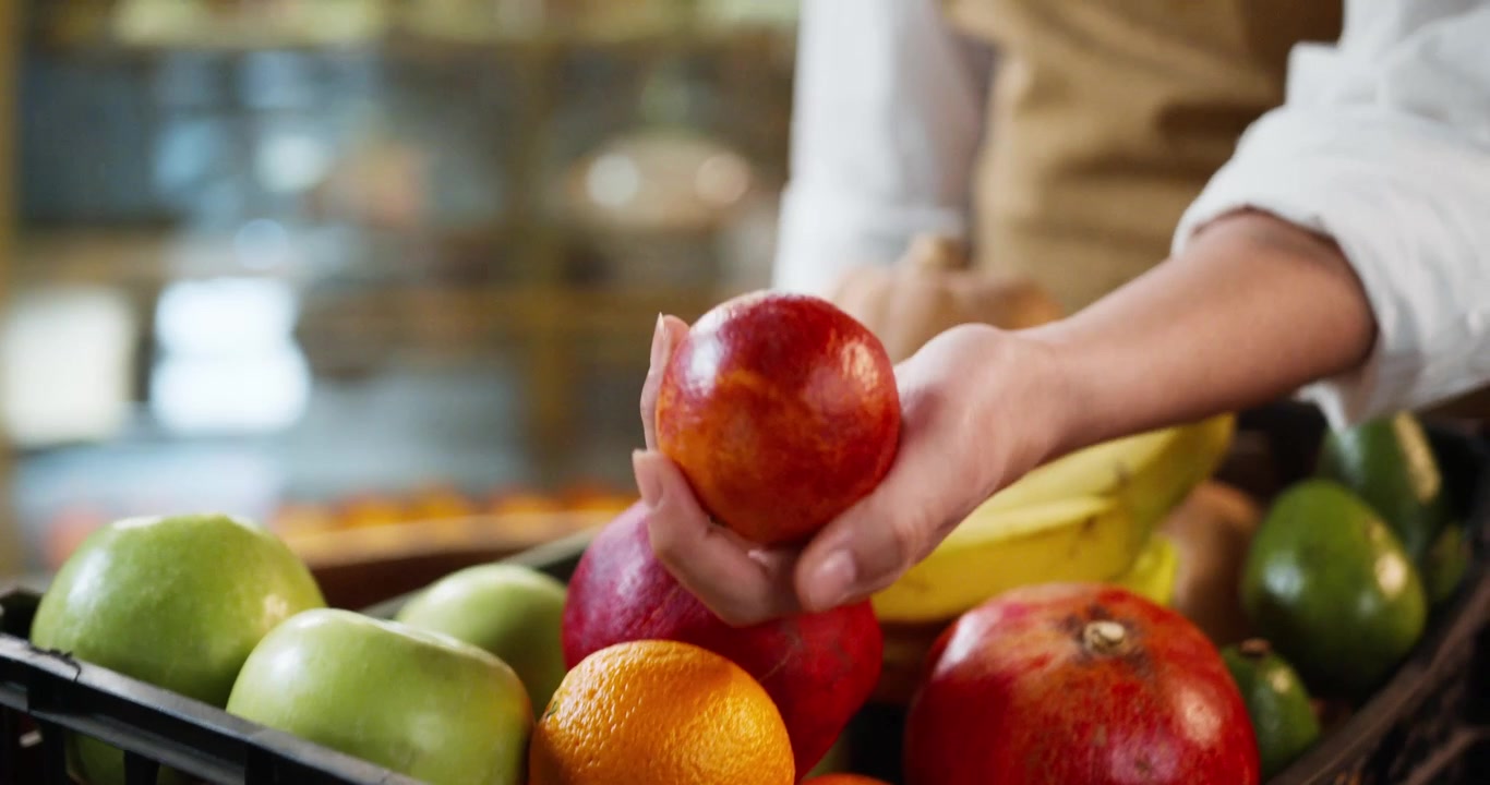 Close-up de mão segurando nectarina fresca sobre uma exibição de frutas variadas com maçãs, bananas, laranjas e romãs. Seleção de produtos saudáveis em supermercado durante o dia.
