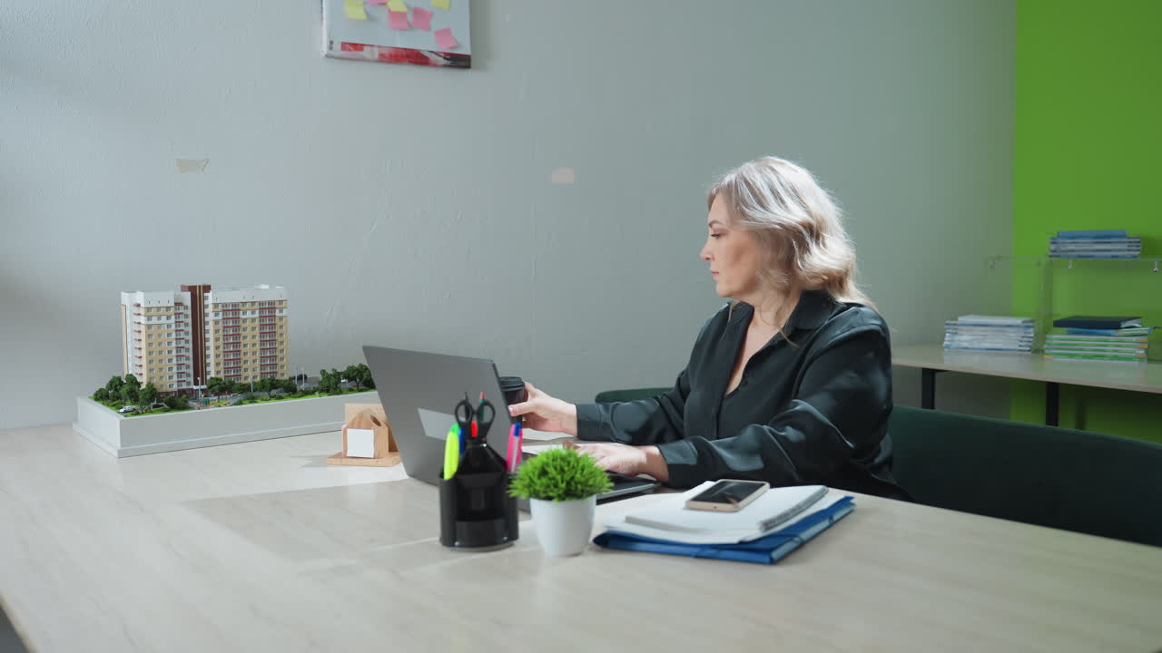 Office assistant typing on laptop in bright workspace pauses momentarily to enjoy coffee before continuing task, surrounded by model buildings, stationery, and organized documents on desk