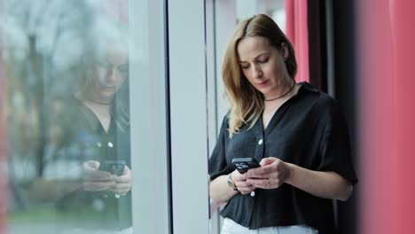 focused woman stands by window texting on smartphone in calm urban silence, riga