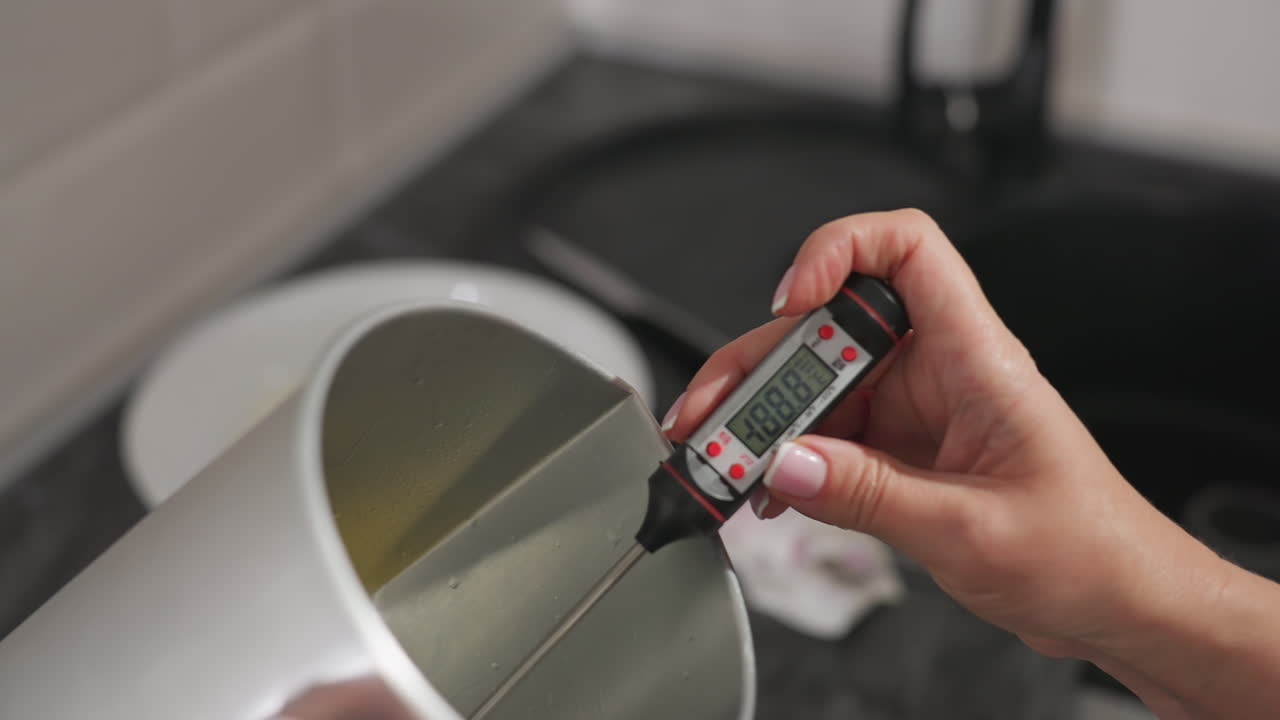 Fair lady switches on digital food thermometer and gently dips it into bowl containing warm mixture in modern kitchen, carefully monitoring temperature during food preparation with full focus