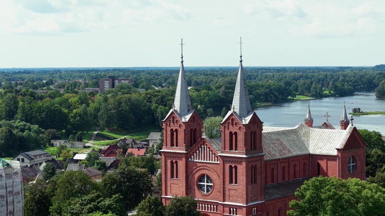 Splendido video drone cattura la Chiesa di San Giovanni Battista a Plungė, Lituania, con torri in mattoni rossi, vegetazione lussureggiante, fiume scintillante e luce diurna chiara in un'ampia inquadratura di apertura