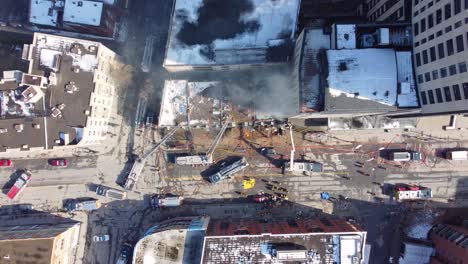 a top down drone shot of black smoke billowing from the rooftop of a building after a devastating fire, on the ground the fired department attempt to contain the smoldering fire