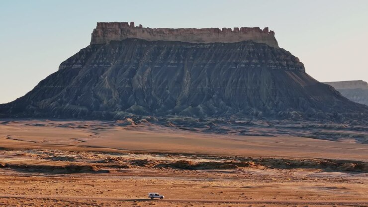 Formazione isolata di mesa desertica che si erge sopra la pianura desertica arida dello Utah, ripresa aerea statica con un camper fuoristrada in movimento.