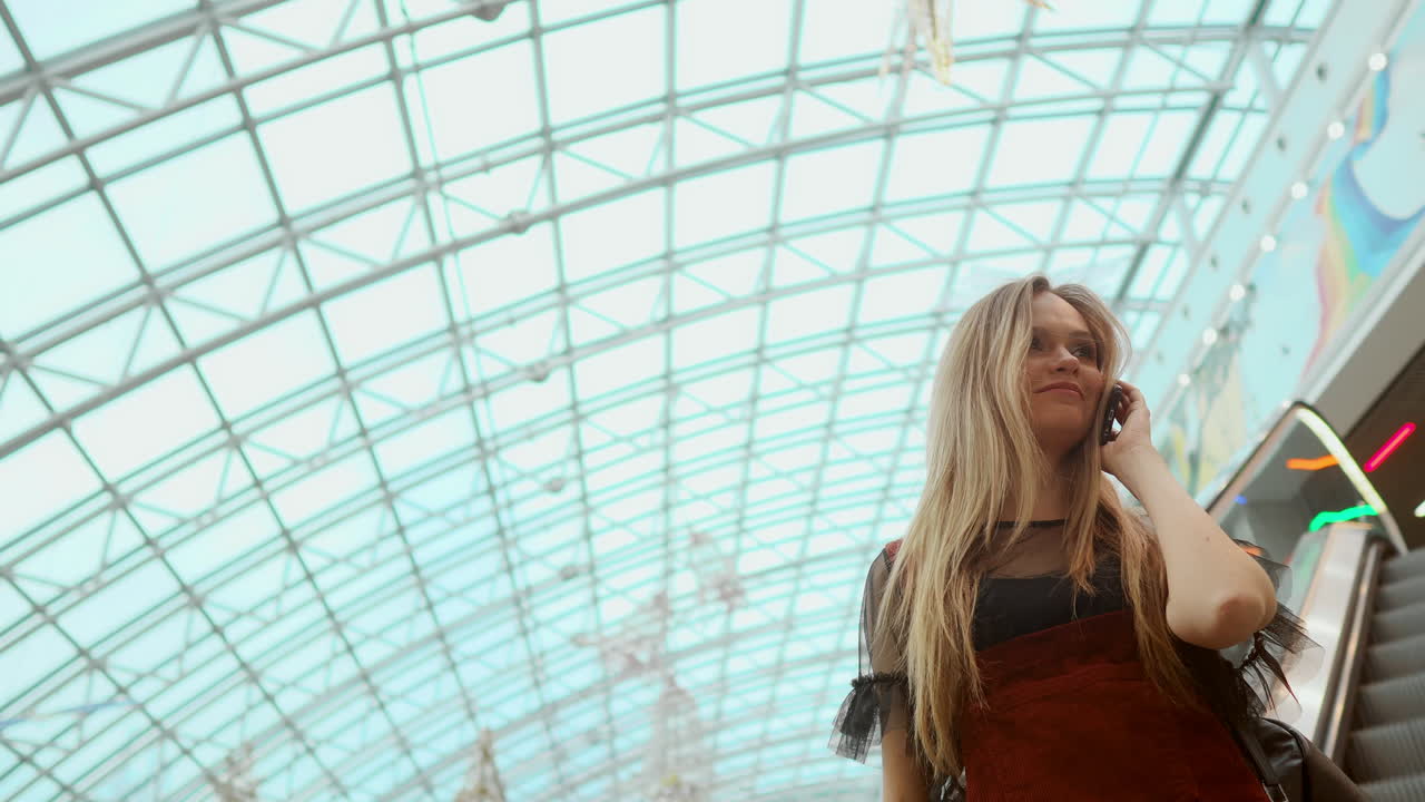una hermosa chica baja por la escalera mecánica y habla por teléfono en el centro comercial.