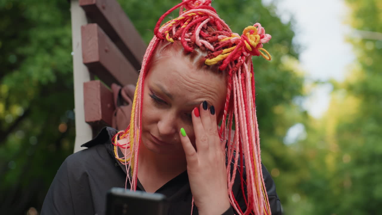 벤치에 앉아 휴대폰을 보며 차분하게 컬러풀한 땋은 머리를 하고 있는 백인 여성의 클로즈업 사진이에요. 체념한 표정이고, 배경에는 멀리 있는 인물이 있으며, 부드러운 공원 조명이 사색적인 분위기를 연출해요.