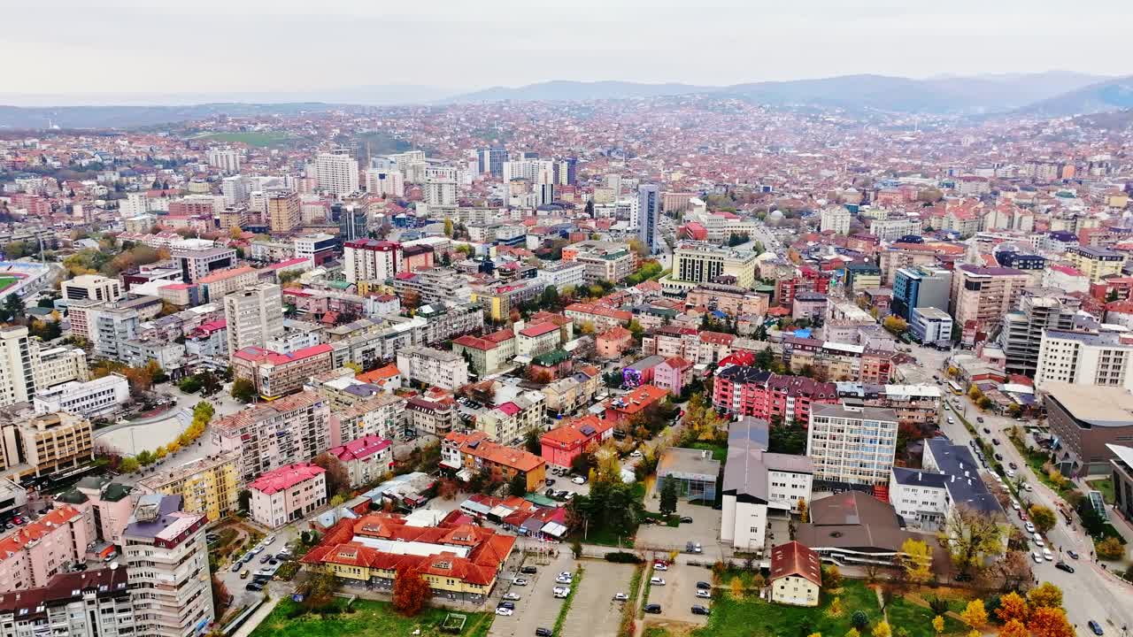 Tripod-style drone, autumn colors across Pristina urban sprawl under grey skies