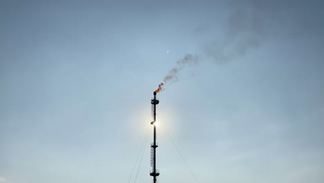 Sun-shining-behind-smoking-refinery-gas-flare-tower-against-blue-sky