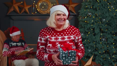 mature grandmother in festive sweater presenting christmas gift box smiling looking at camera