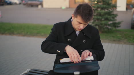 investor seated outdoors after work, going through documents, taking diary from bag in urban setting, office building, street light, and parked car at twilight