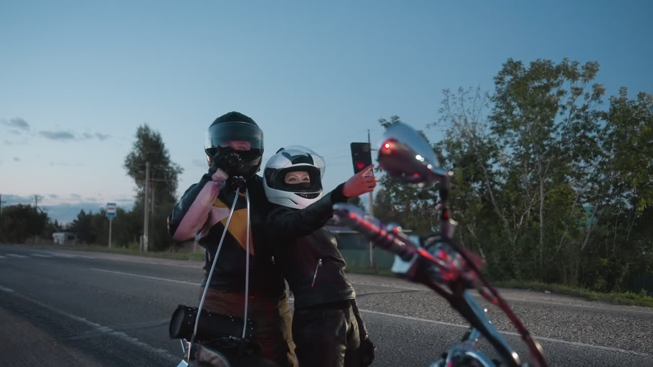 Man wearing motorcycle helmet adjusts visor while standing with woman in helmet as she holds smartphone taking picture of them on roadside during evening with soft fading light and passing headlights