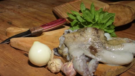 raw octopus on wooden cutting board with onion, garlic, herbs, knife