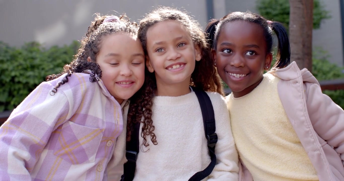 African-American schoolgirls moving closer left girl leaning reaching posing with backpack on bench