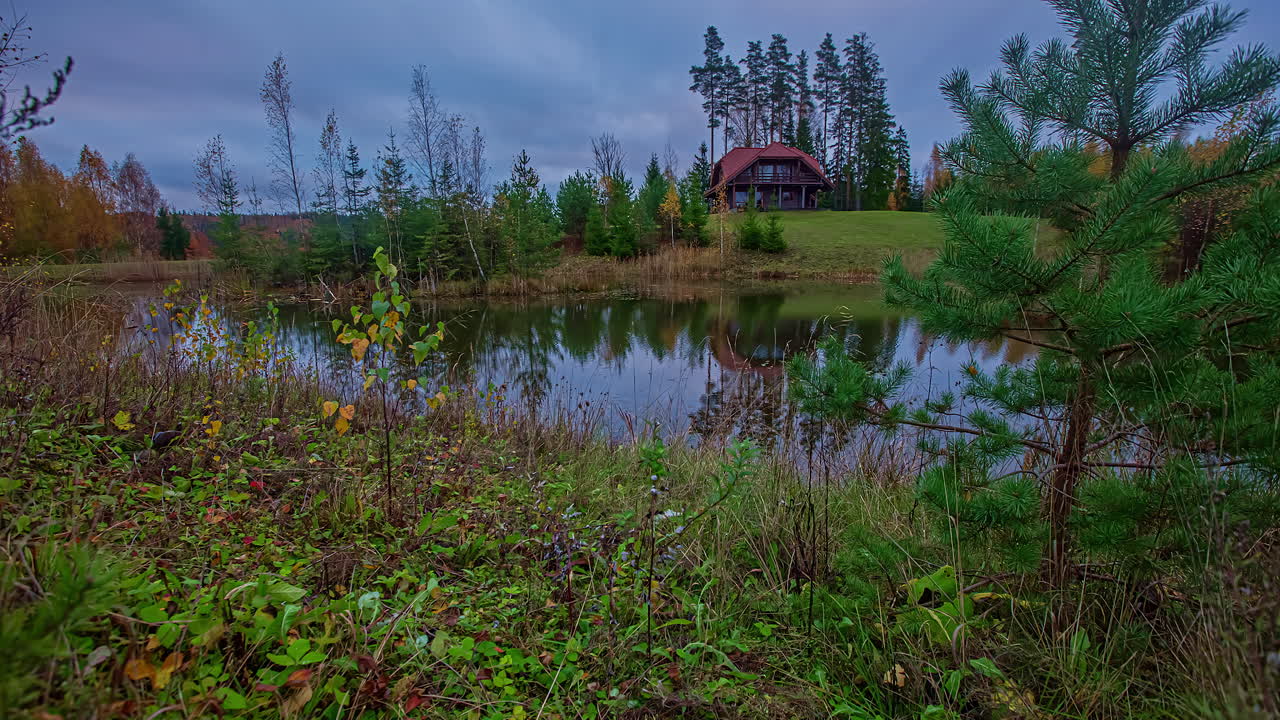 Timelapse-optagelse af et træsommerhus i baggrunden med udsigt over en sø i forgrunden på en overskyet aften