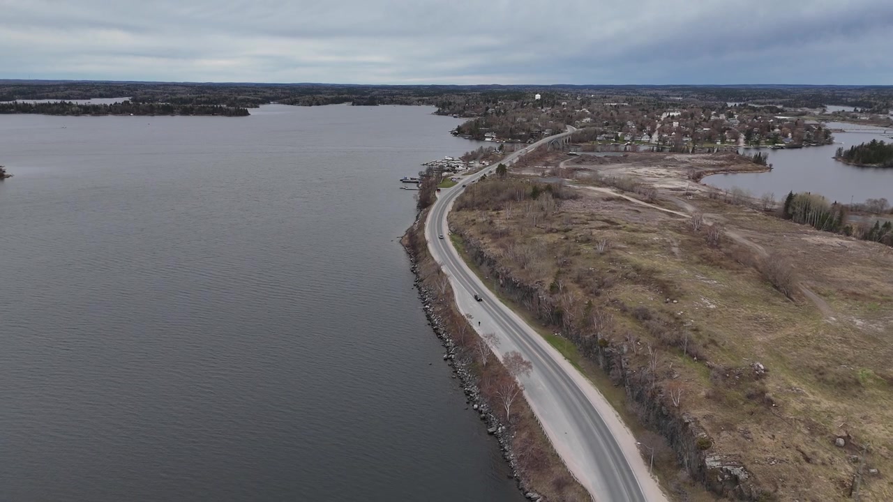 Lago dei Boschi vicino a Kenora, Ontario. Ampia ripresa aerea di una strada costiera con auto che viaggiano lungo il bordo dell'acqua, circondata da foresta e lago sotto un cielo primaverile nuvoloso