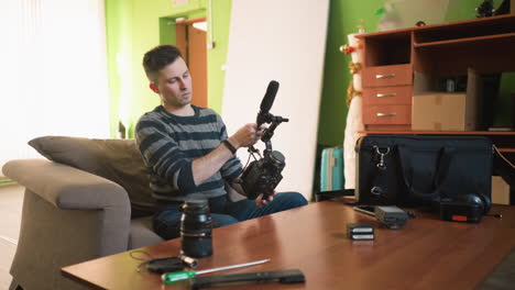 man reaches for camera gear in home studio, organizing equipment on wooden table. visible camera lenses, microphone, batteries, and other filming tools. cozy workspace with green walls