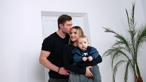 loving man kisses his wife on the head. blond woman holding her little infant baby in hands. family portrait.