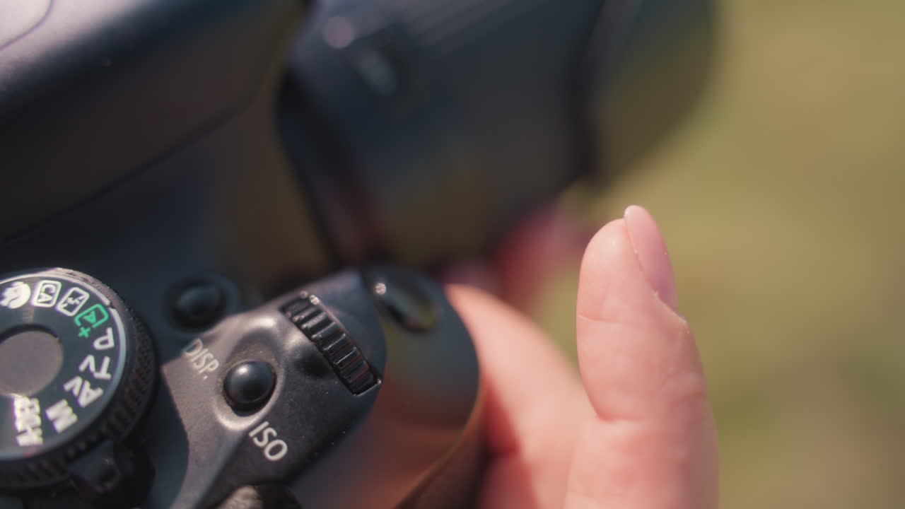 Close up of fair skin finger adjusting camera dial during outdoor shoot, with lens and blurry grassy field in background under sunlight, capturing technical detail of camera operation moment
