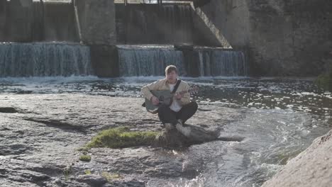 young man playing guitar sitting on the bank of a mountain river on a background of rocks. concept of freedom relaxation. place