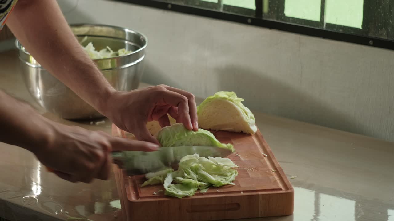 Hands slicing cabbage vegetables placing into metal bowl on kitchen counter indoors in soft light, cooking lifestyle detail, static