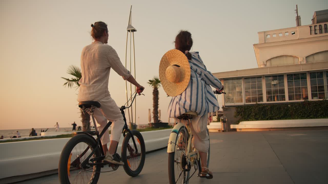 vista trasera de un chico feliz con ropa blanca en una bicicleta negra y una chica con un sombrero de paja en un paseo en bicicleta a lo largo de la playa cerca del mar al amanecer en verano. ocio activo y cita en movimiento en bicicletas cerca del mar en la playa