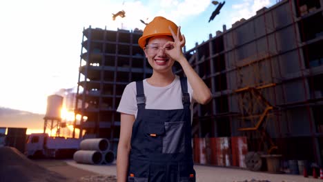 trabajadora asiática con gafas de protección y casco de seguridad sonriendo y mostrando un gesto de acuerdo a la cámara mientras está de pie en el sitio de construcción