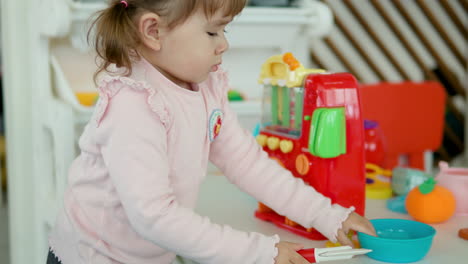 Niña-Pequeña-Con-Coletas-Jugando-Con-Utensilios-De-Cocina-Juguetes-Coloridos-De-Plástico-En-Casa-Fingiendo-Cocinar---Tiro-Medio