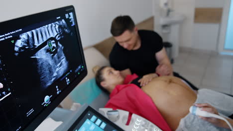 ultrasound machine screen showing the result of pregnancy check up. caring man kissing his expecting wife at backdrop in blur. top view.