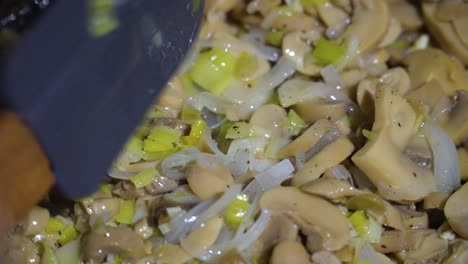 close up of chopped mushrooms, leek and onion in frying pan stirred with spatula