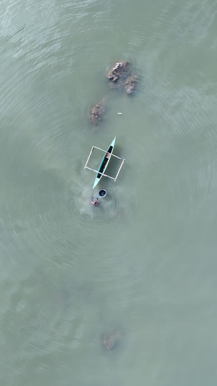 Aerial drone view of a man swimming near a traditional boat with a floating basket in shallow water in the Philippines. Vertical