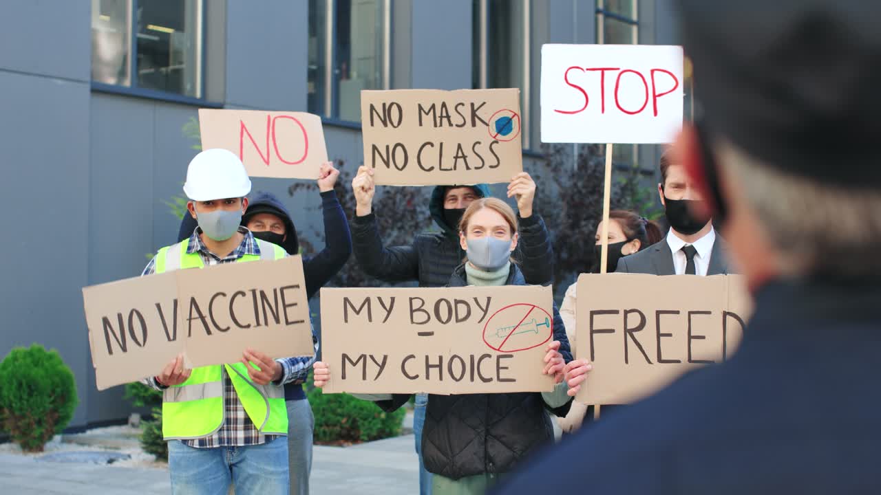 거리에서 코비드 19에 반대하는 시위에서 간판을 들고 고함을 지르는 사람들을 바라보는 경찰관의 뒷모습