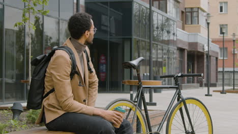 Lächelnder-Amerikanischer-Mann-In-Formeller-Kleidung,-Der-Kaffee-Trinkt,-Während-Er-Neben-Seinem-Fahrrad-Auf-Einer-Holzbank-Auf-Der-Straße-Sitzt