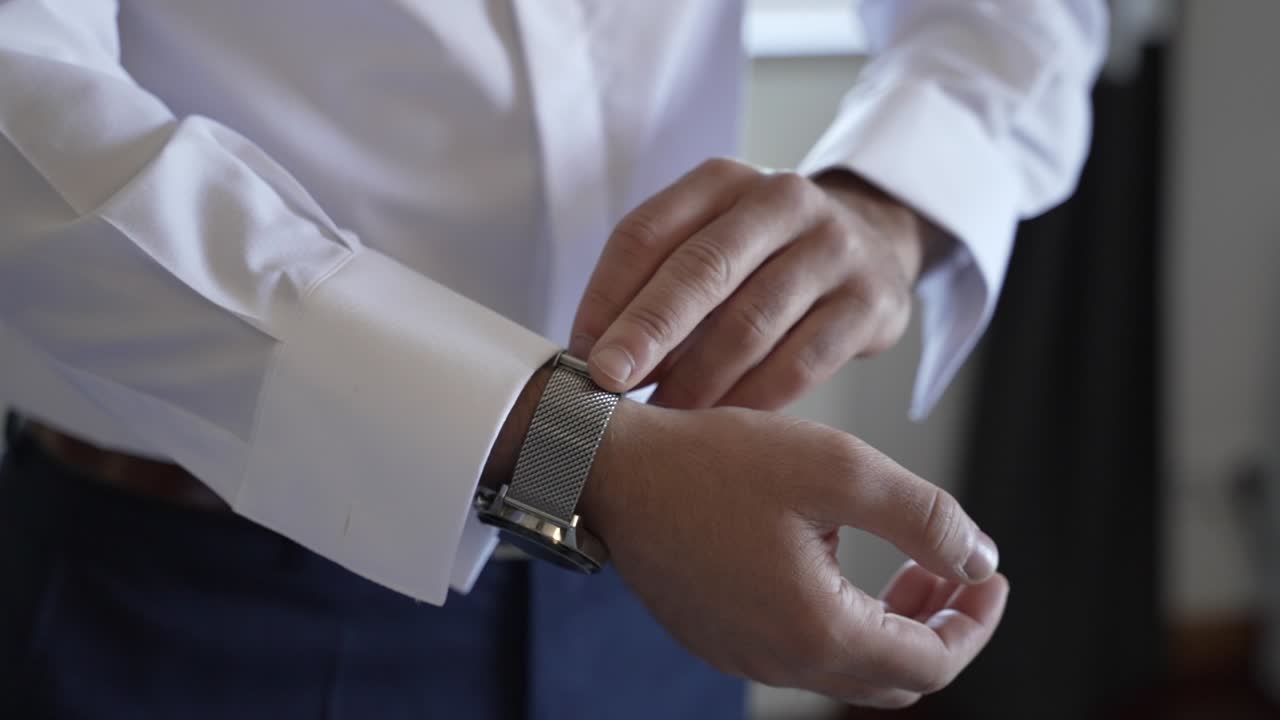 hombre ajustando un reloj de muñeca de acero inoxidable en su mano izquierda mientras lleva una camisa blanca, preparándose para un evento formal