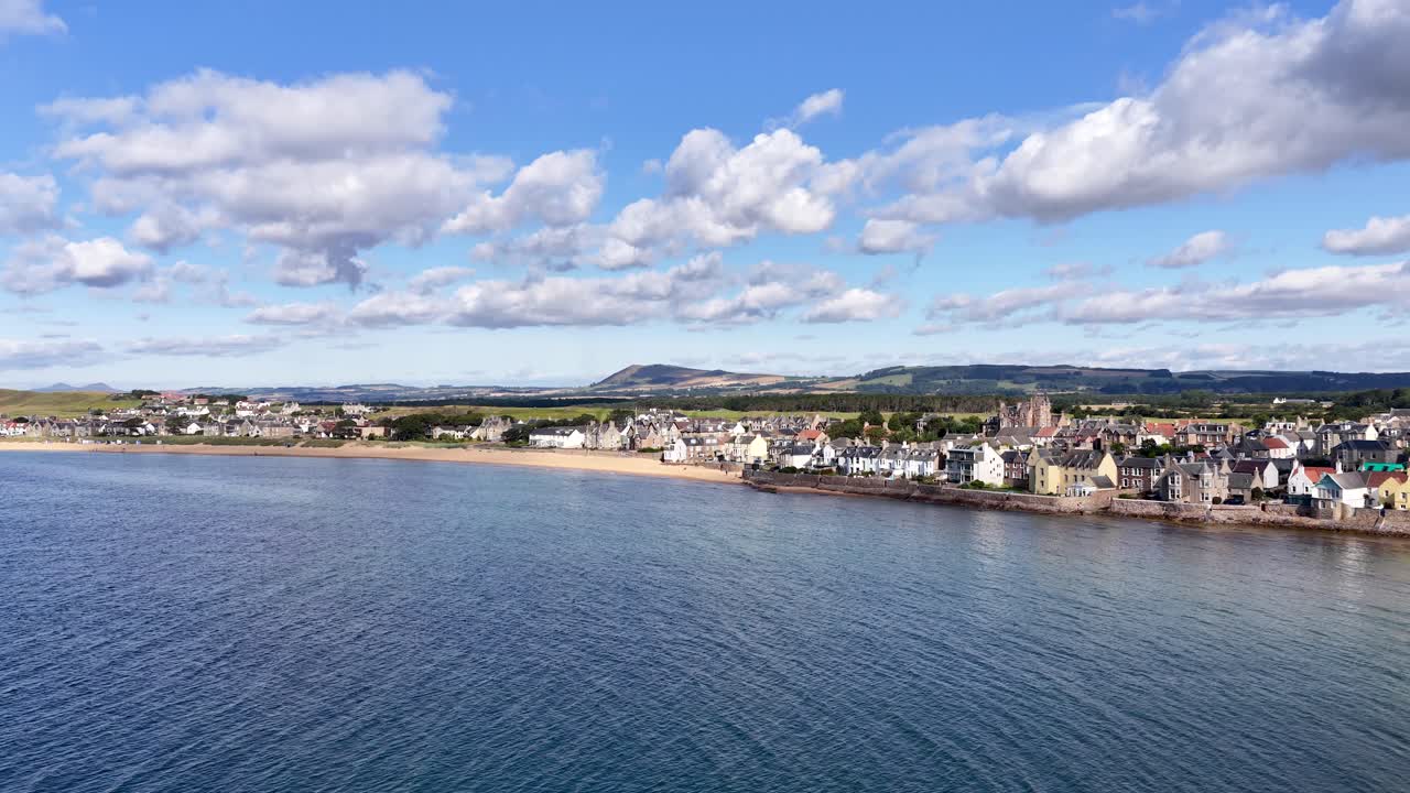 Drone pans over harbor with boats, waterfront village, and scenic landscape under bright daylight