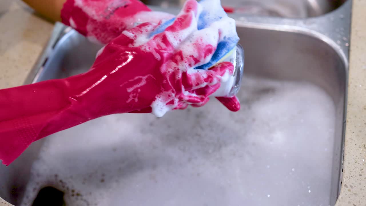 Hands in red gloves meticulously wash a glass with a sponge in a kitchen sink, emphasizing cleanliness