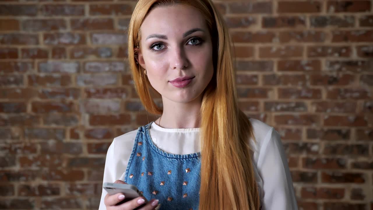 Young alluring blond girl is reading message on smartphone, watching at camera, brick background, communication concept