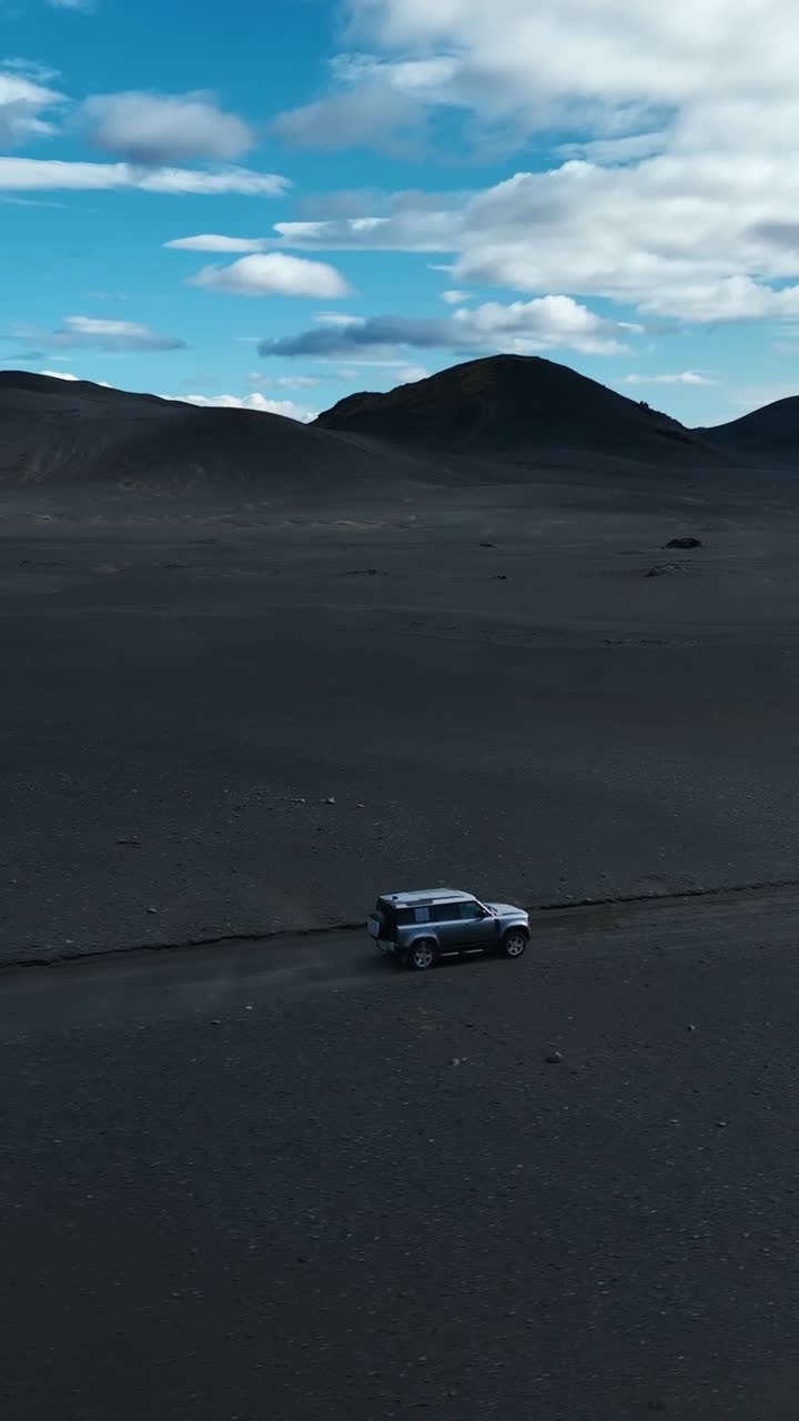 Un fuoristrada 4x4 solitario attraversa un vasto deserto vulcanico in Islanda, circondato da sabbia nera e montagne distanti sotto un cielo parzialmente nuvoloso. Aspra bellezza degli altopiani islandesi.