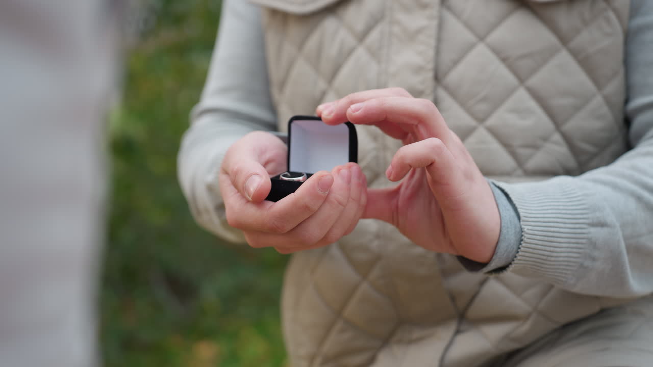 Close up of white man on one knee gently holding ring box in both hands preparing for proposal with blurred background of soft greenery and swaying leaves in romantic calm outdoor atmosphere
