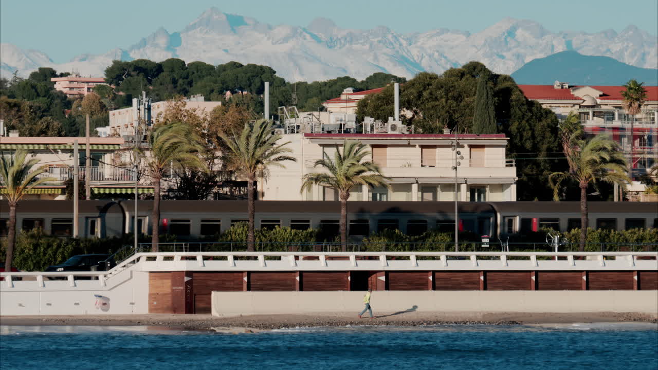 Train moving in front of buildings and palm trees on the shore with a view of the mountains in the background in Antibes, France